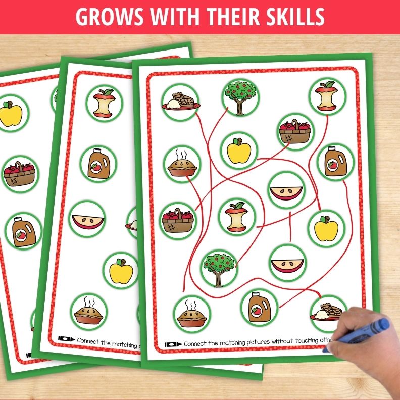 Child's hand holding blue crayon over apple-themed line tracing worksheet with matching pictures connected by red lines