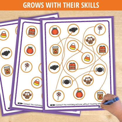 Child's hand drawing lines connecting Halloween pictures like pumpkins, bats, and candy corn in matching activity worksheet.